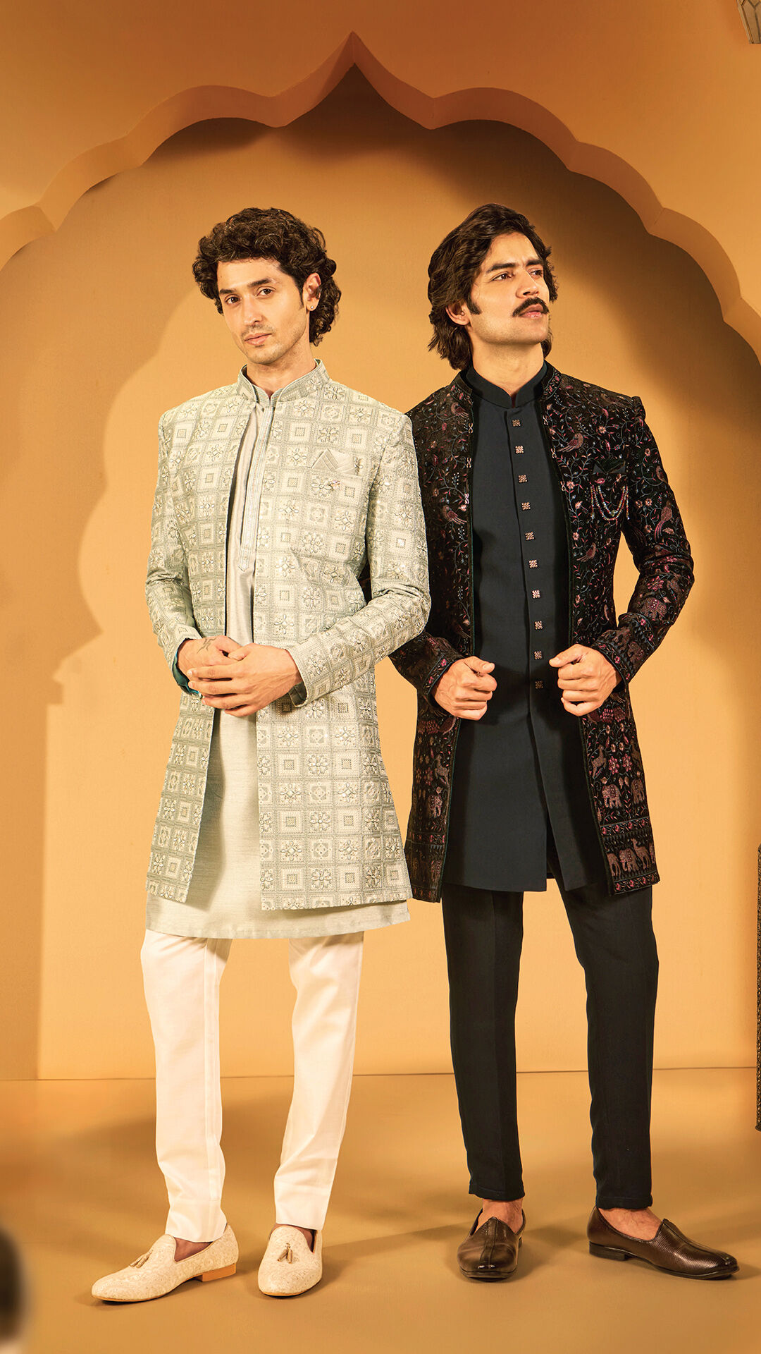 Two men in modern wedding outfits—one in a mint green printed sherwani and the other in a black embroidered Indo-western jacket